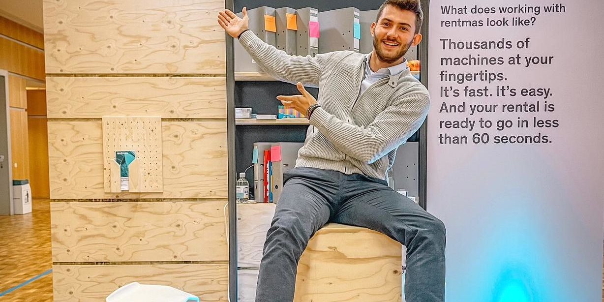 Giacomo at rentmas Bauma booth 2022 Partner care guru Giacomo sitting on a wooden table at Bauma, drawing attention to the rentmas sign behind him.