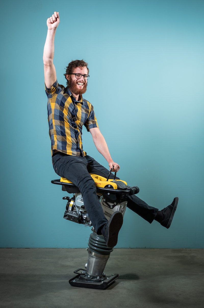 A smiling man riding a piece of construction equipment like a rodeo bull. His right hand is held up in the air.